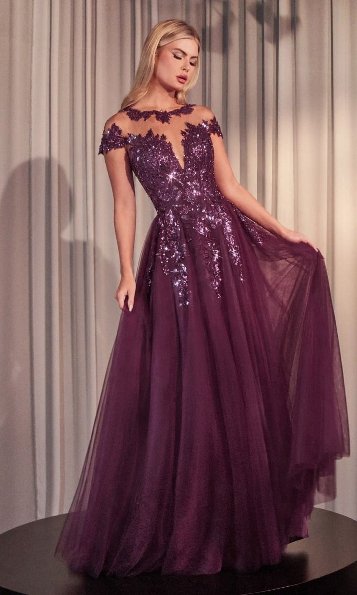 A woman in the Andrea and Leo A1475 long prom dress in eggplant, featuring an off-the-shoulder sequined bodice and tulle A-line skirt, stands before light curtains.
